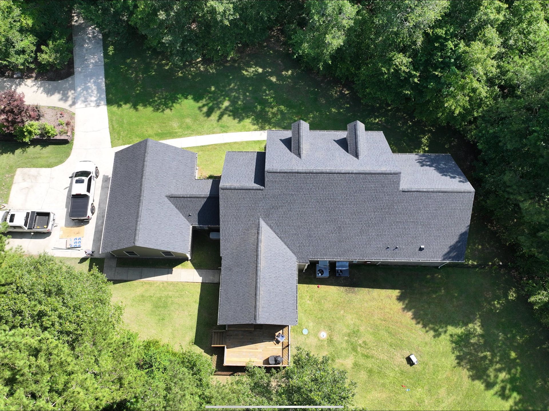 Overhead view of a house with a dark roof, surrounded by green grass and trees, and a driveway with parked cars.