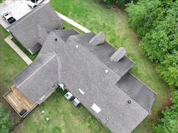 Overhead view of a house with a gray shingle roof, surrounded by green grass and trees.