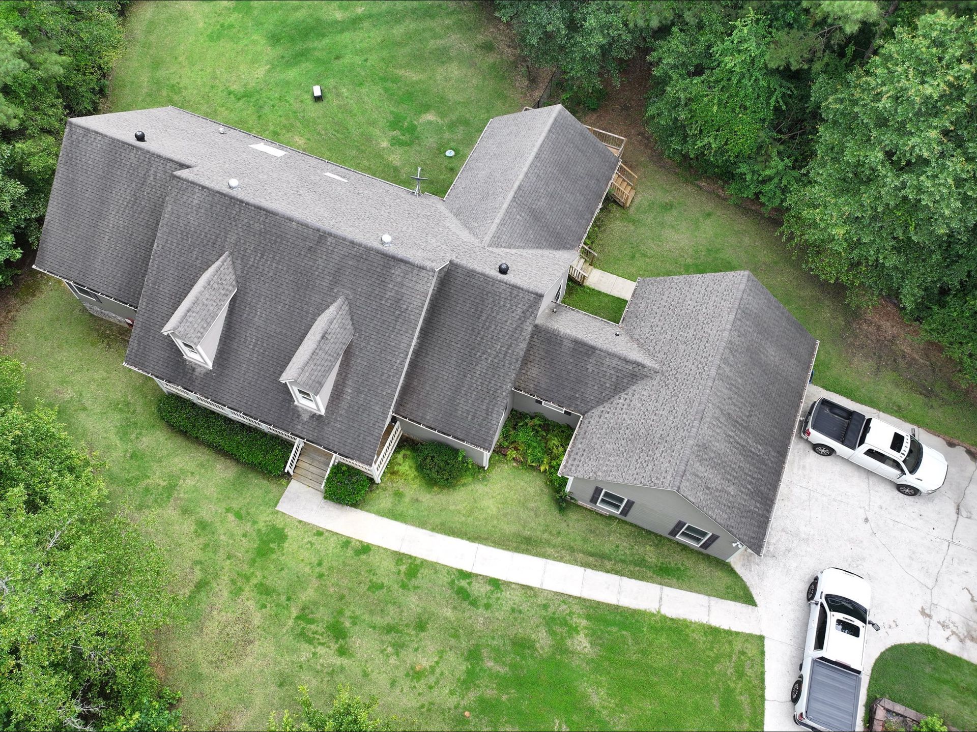 Overhead view of a house with a gray roof, dormers, and two white vehicles parked on a gravel driveway.