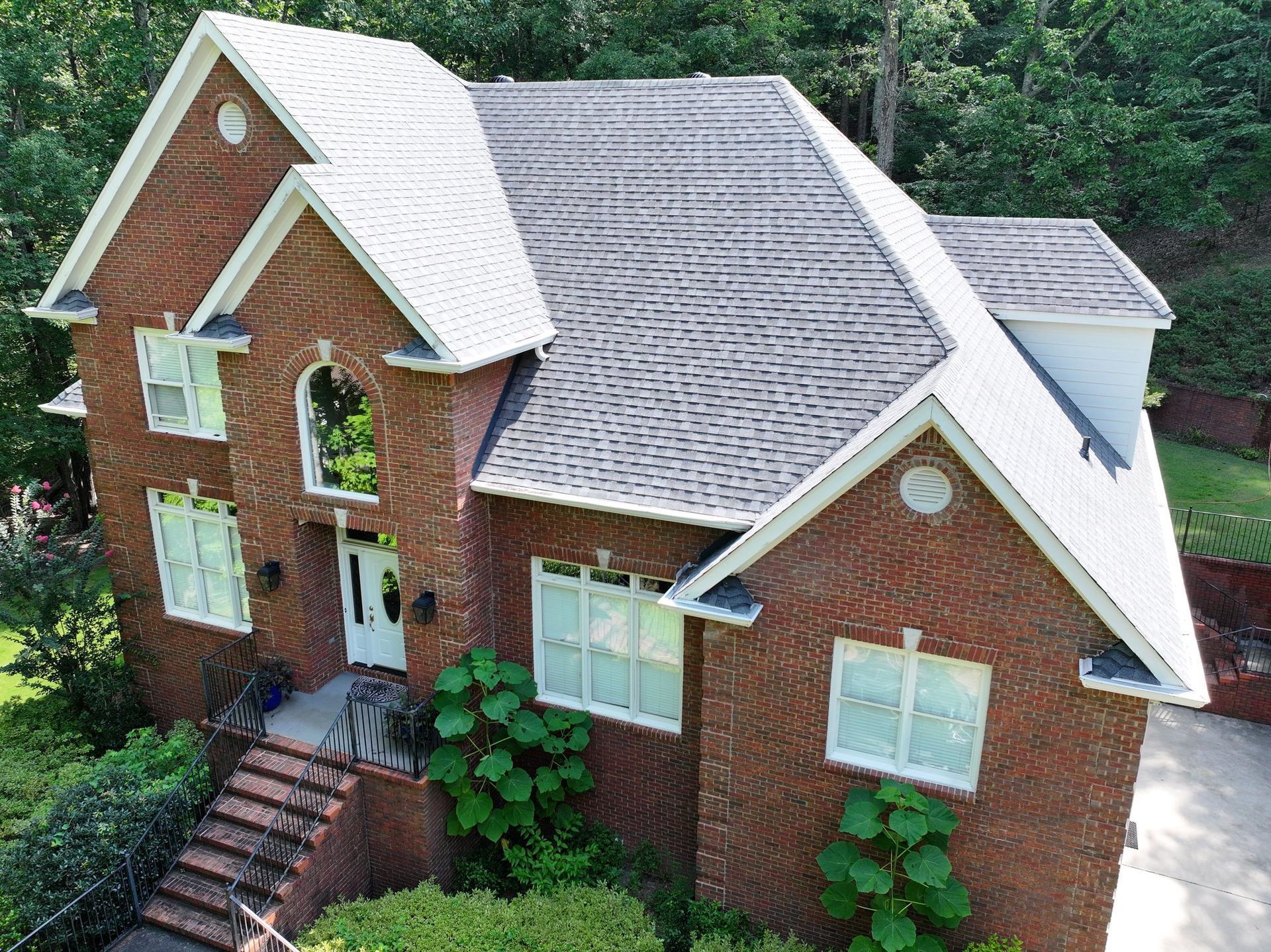 Two-story brick house with a gray roof and white trim, surrounded by greenery.