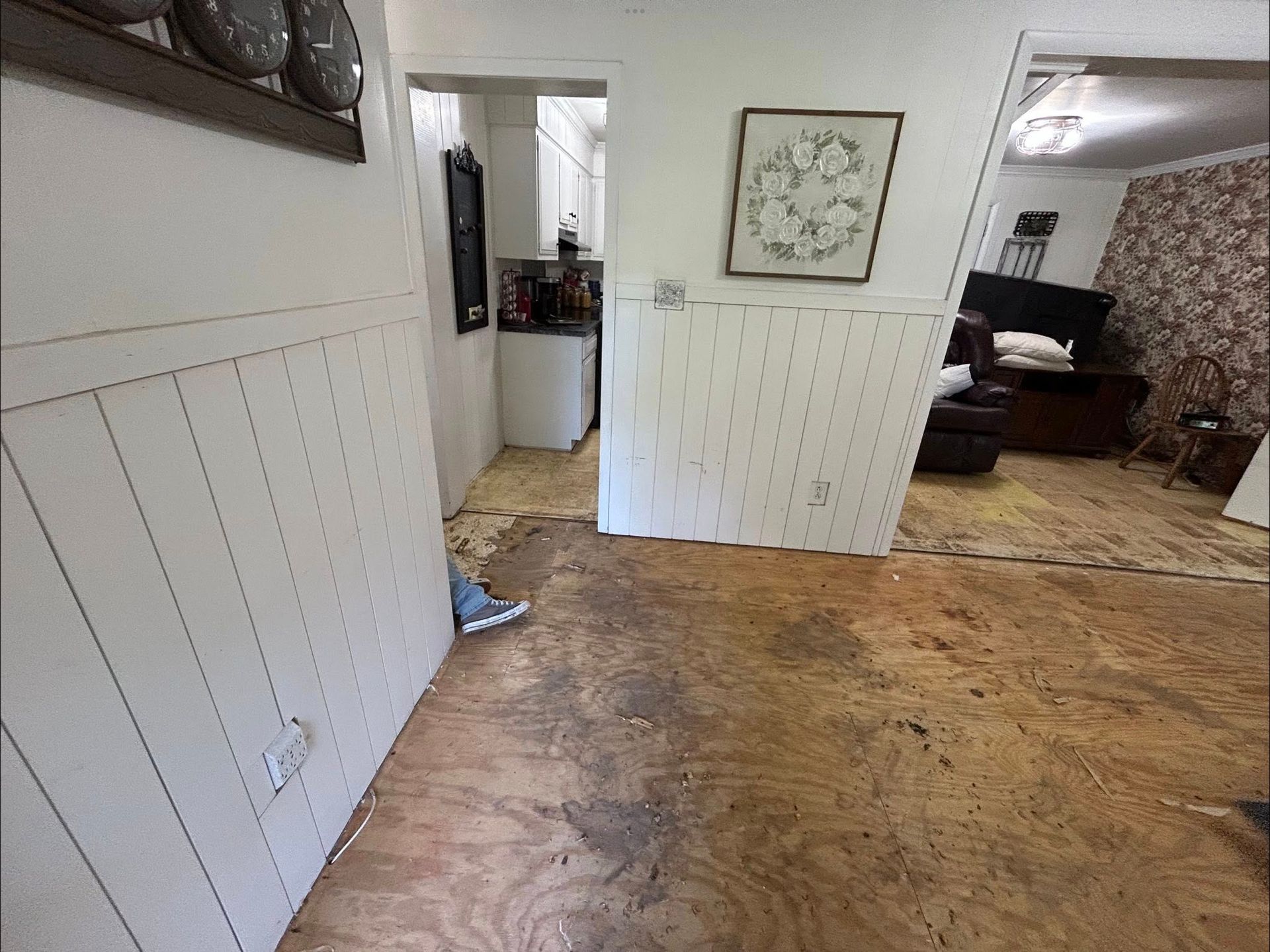 Interior view of a home with exposed subfloor. White walls and a doorway lead to a kitchen and living area.