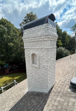White brick chimney on a shingled roof, topped with a black cap, set against a backdrop of trees and a blue sky.