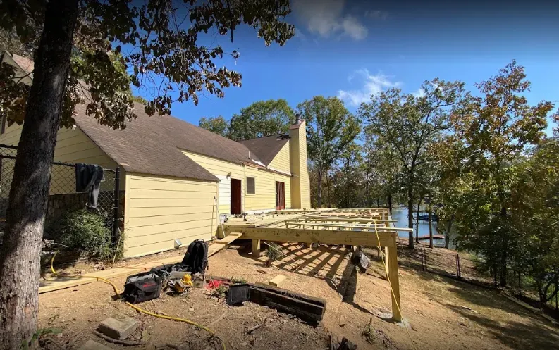 Construction of a wooden deck next to a yellow house with trees, overlooking a lake on a sunny day.