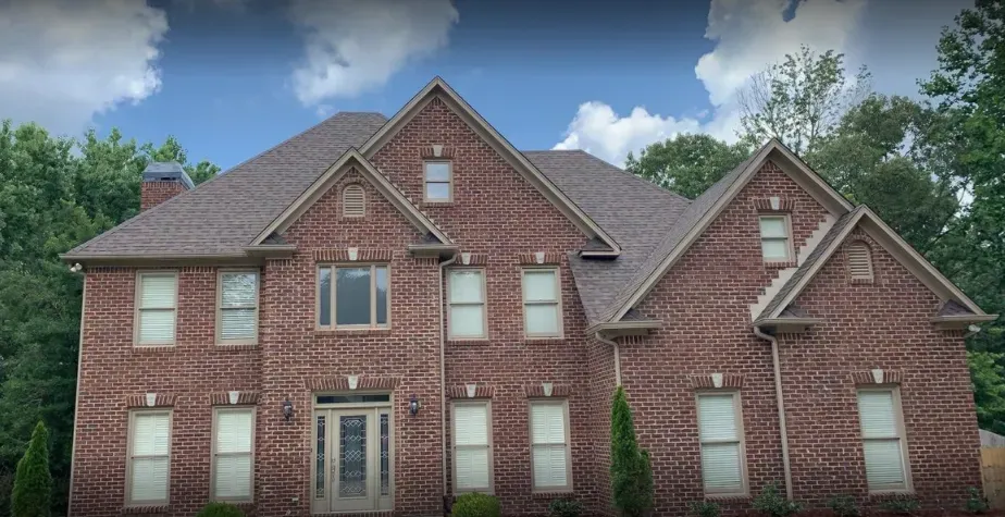 Brick two-story house with a brown roof and white-framed windows against a partly cloudy sky.