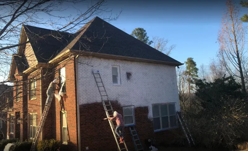 Workers painting the exterior of a two-story brick and white house, using ladders.