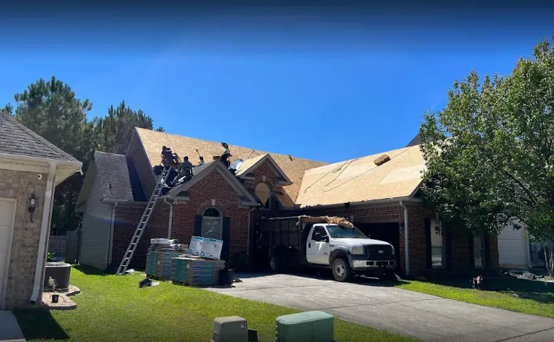 Roofers working on a residential home with a truck and materials on the driveway under a blue sky.
