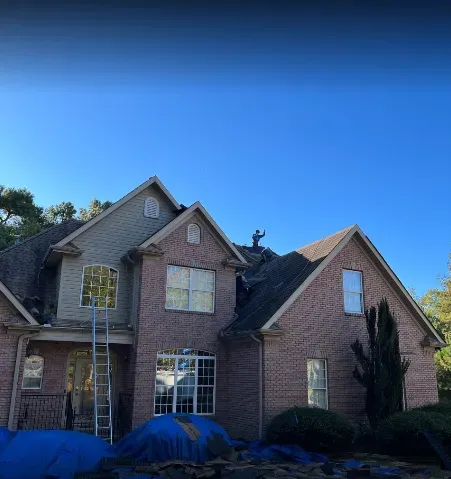 Residential house with a person on the roof; construction underway. Blue tarps and a ladder present.