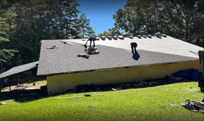 Two people on a roof replacing shingles, surrounded by trees.