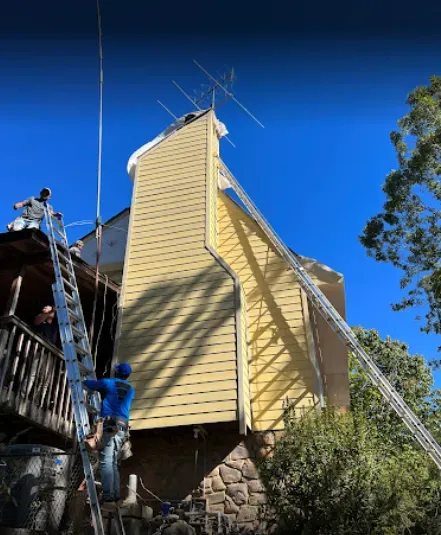 Workers on ladders near a tall yellow chimney with an antenna against a blue sky.