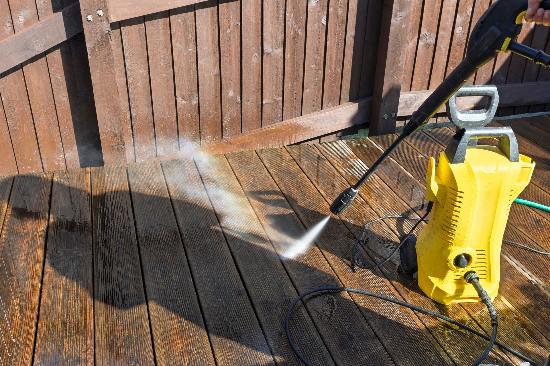 A yellow pressure washer spraying water on a wooden deck, cleaning it. Sunlight casts a shadow.