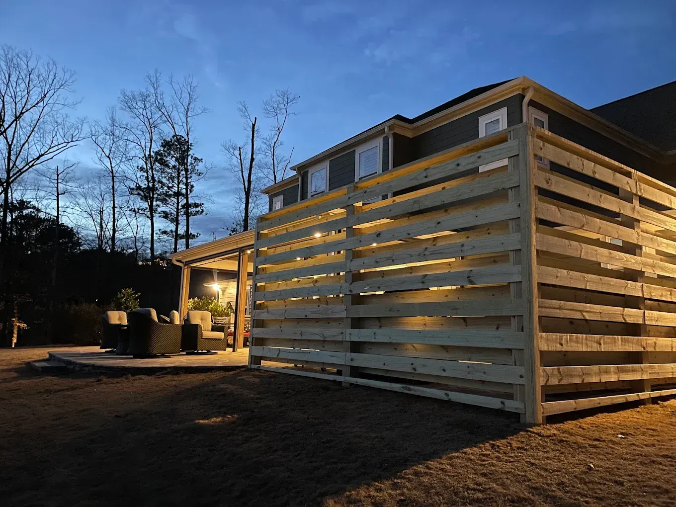 Wooden privacy fence next to a house with a lit patio and lawn at dusk.