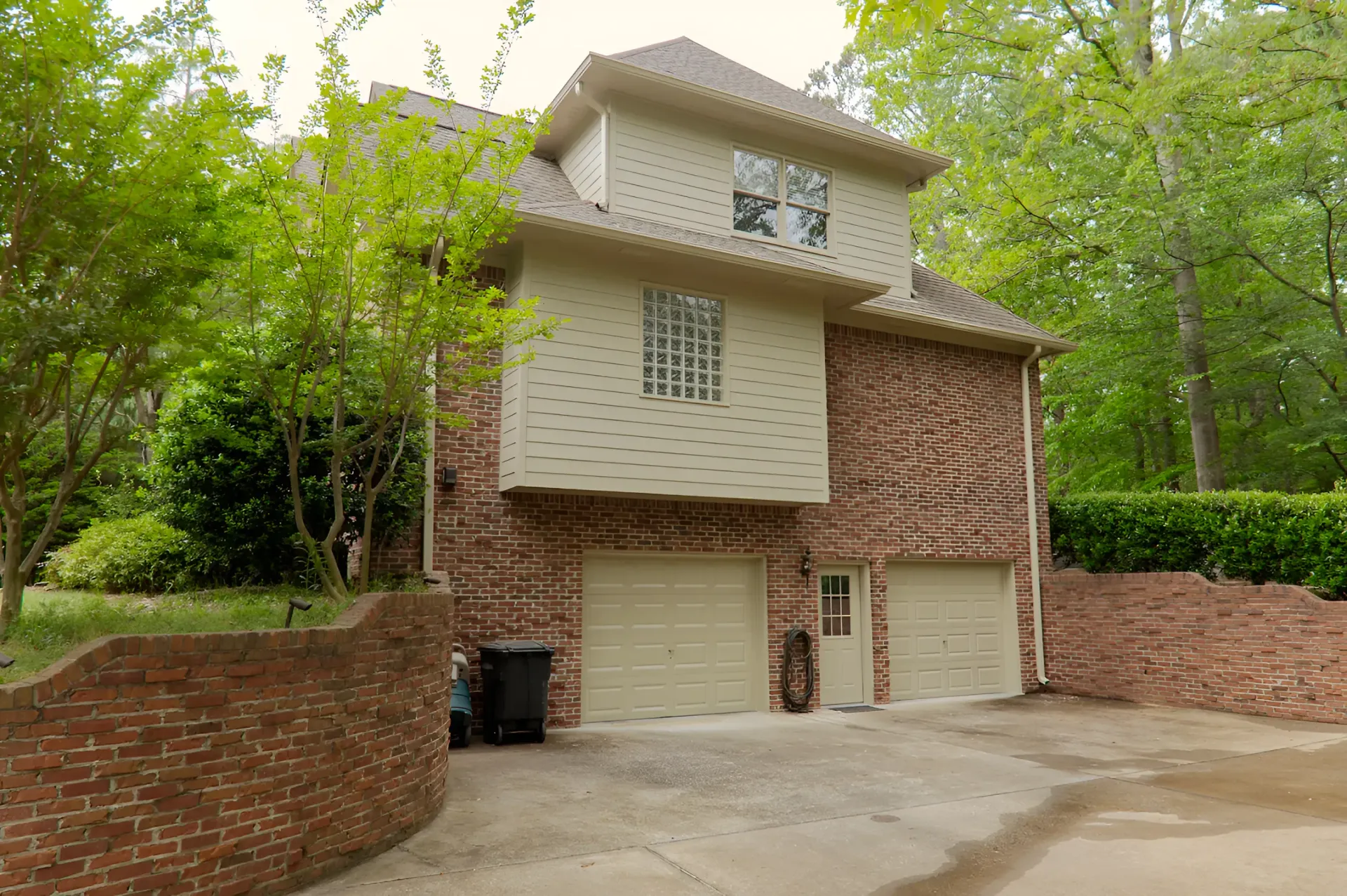 Two-story house with brick and beige siding, garage doors, and a small front yard surrounded by trees.