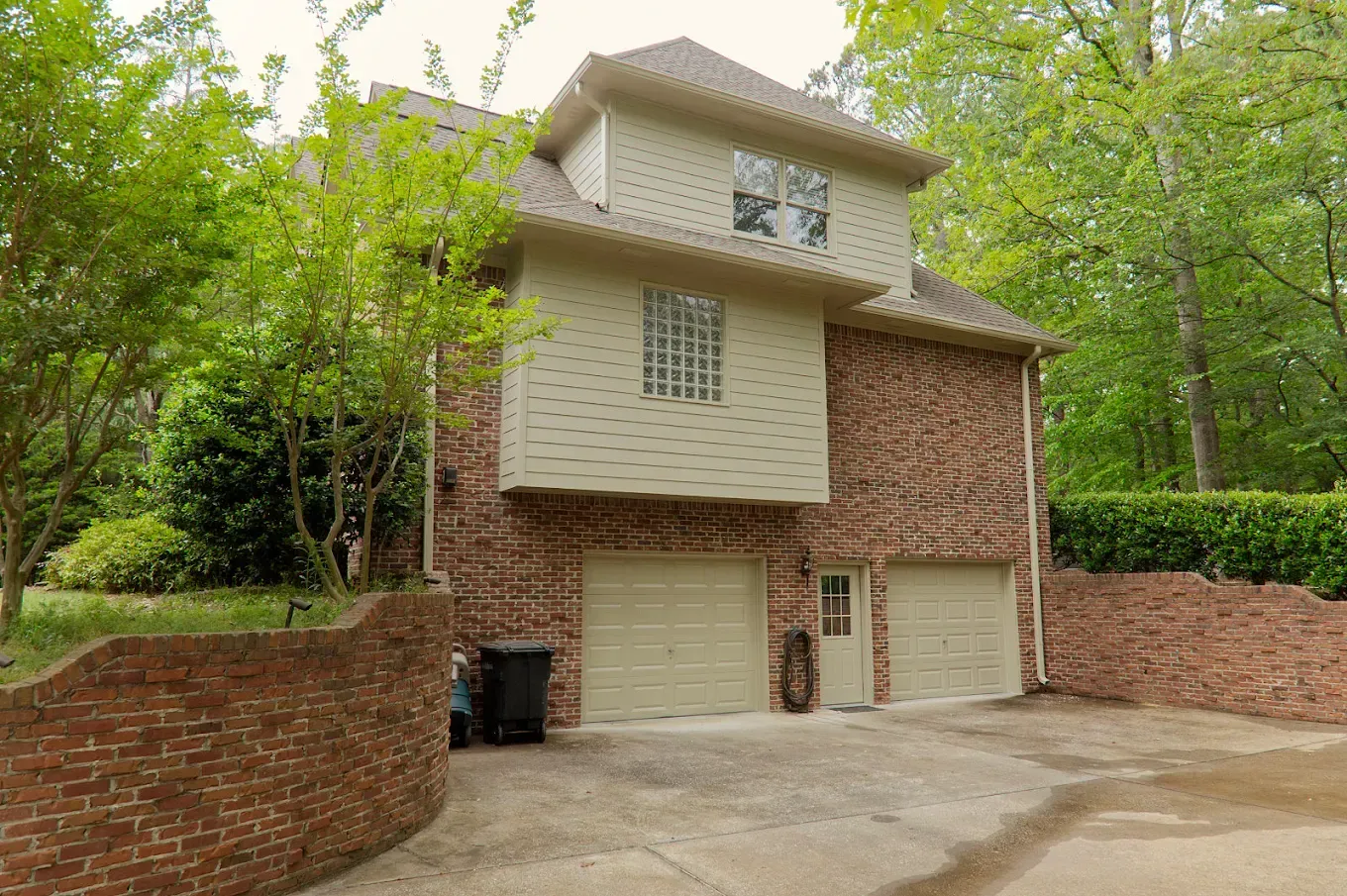 Two-story brick house with a garage and tan siding. Trees and a brick wall surround the home.