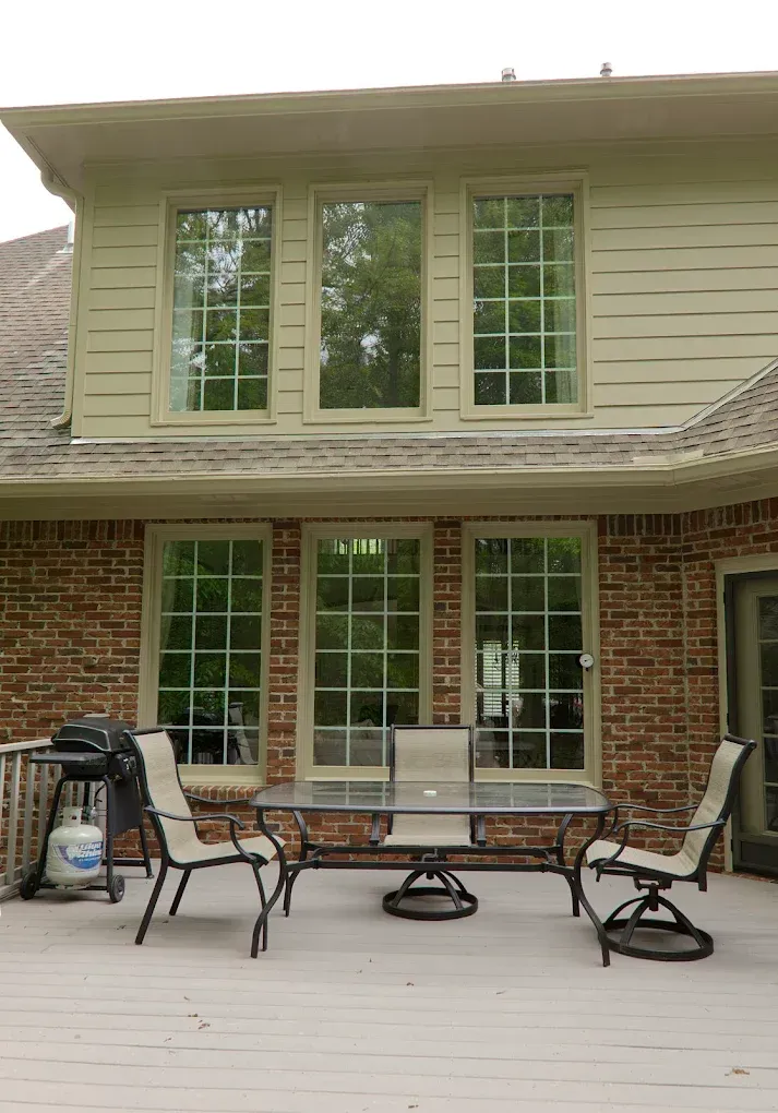 Backyard deck with brick and siding house, outdoor dining set, grill, tall windows.