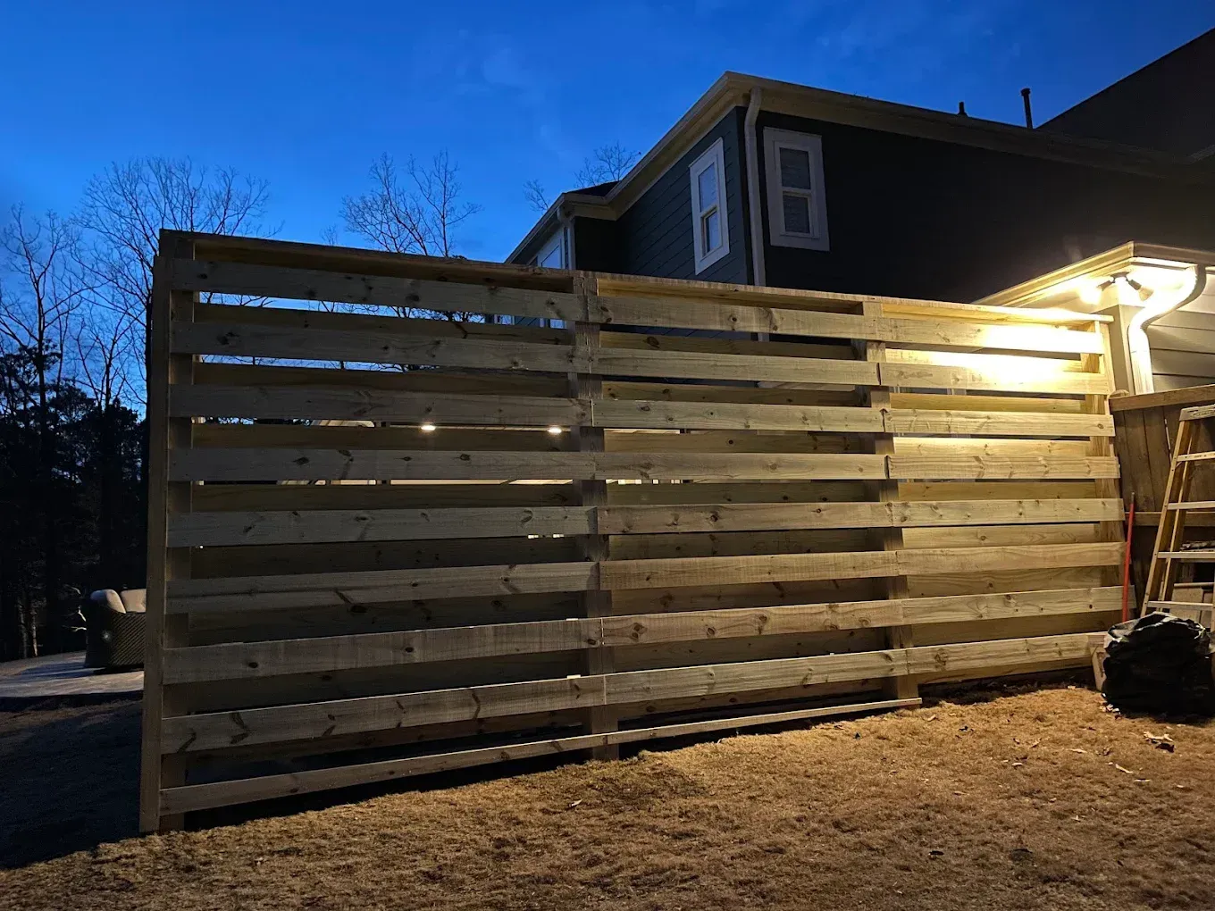 Wooden pallet fence in front of a house at dusk, lit by outdoor lights.