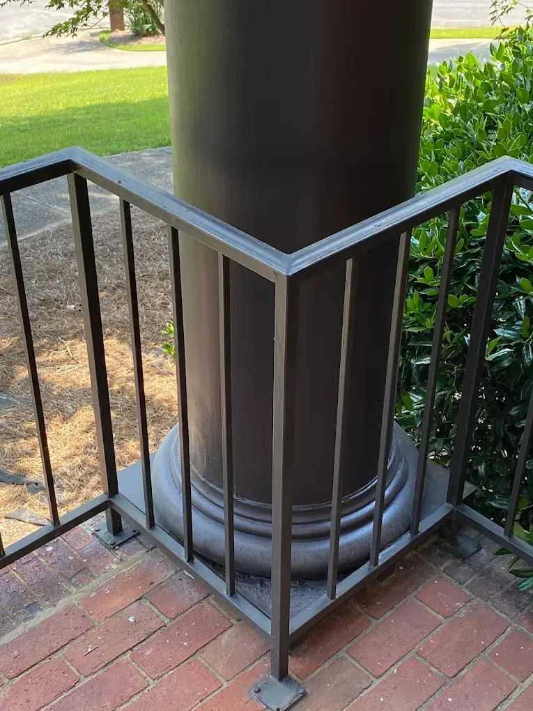 Black metal railing around a dark column on a brick surface, next to greenery.