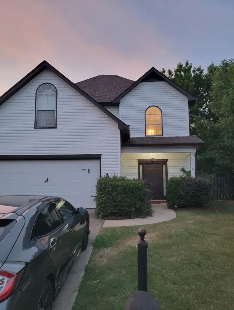 Two-story white house with brown roof and a car parked in front. Evening sky in the background.