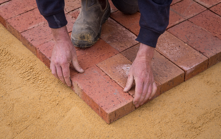 A man is laying bricks on a sandy surface.