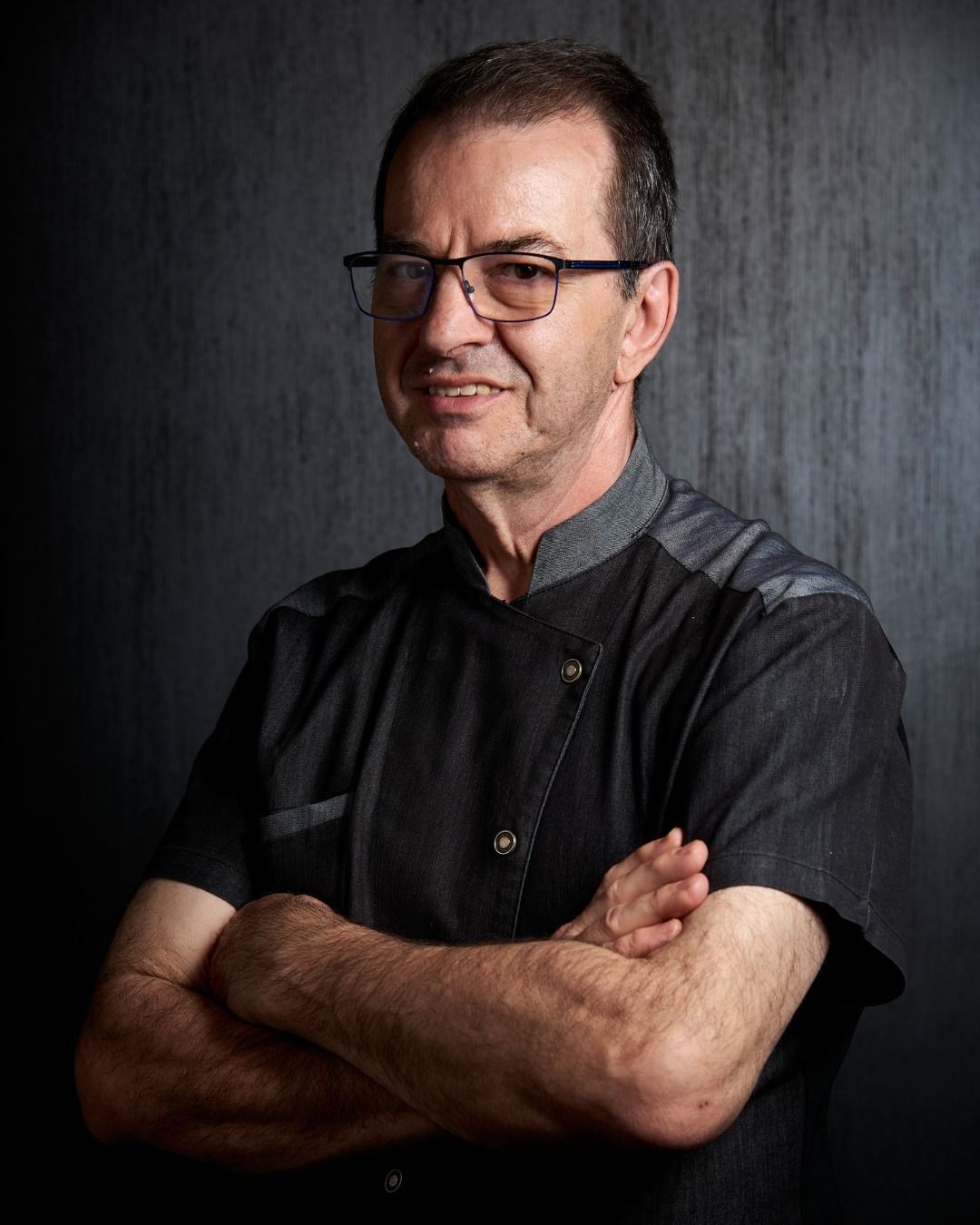 Un uomo che indossa occhiali e giacca da cuoco è in piedi con le braccia incrociate.