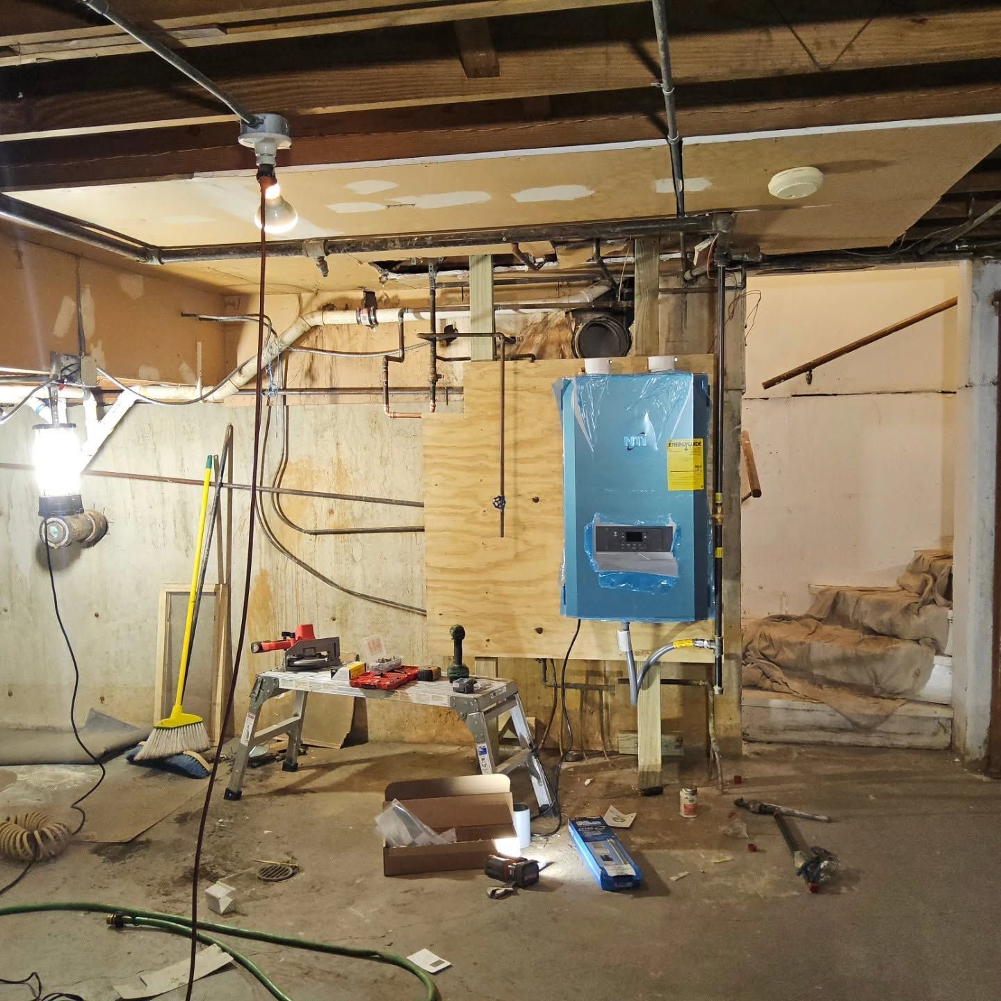 Basement under renovation with exposed joists, hanging wires, tools, and a blue boiler unit.