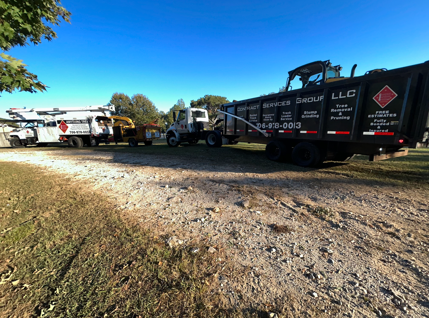 Several work trucks parked on gravel, including a dumpster truck with a black container. Clear blue sky.