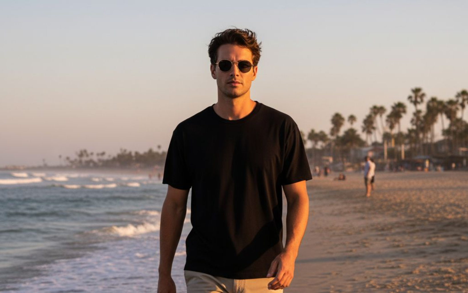 Man in black t-shirt and sunglasses walks on beach at sunset, ocean waves visible.