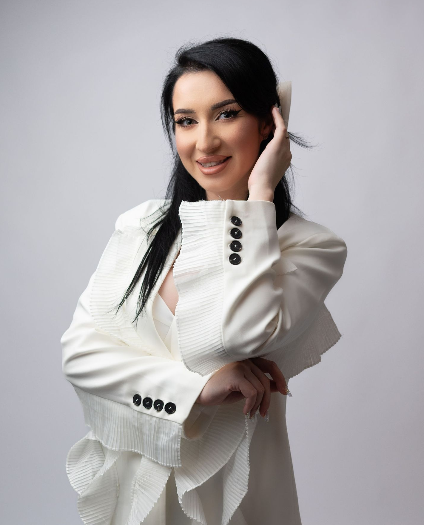 Woman in white blazer smiles, hand in hair, against a light background.
