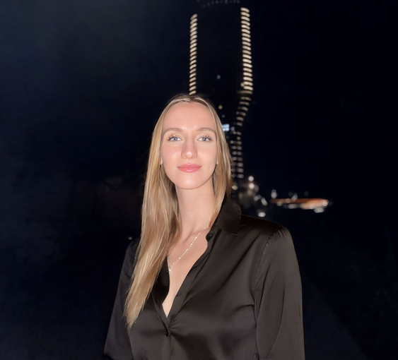 Woman with long blonde hair in a black button-up shirt, standing outside at night with a lit-up structure in the background.