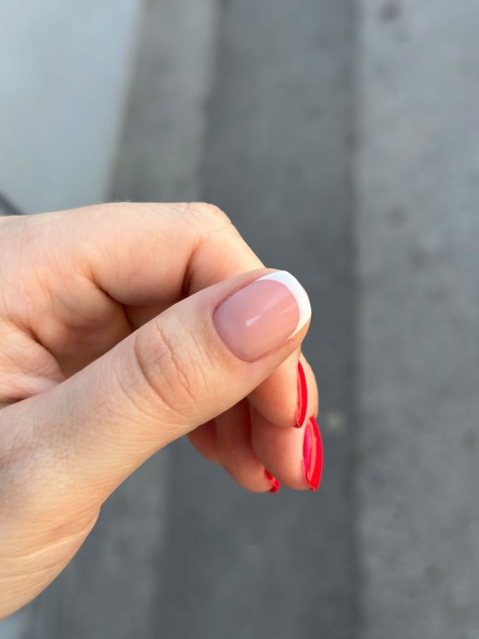 Hand with French manicure on one nail and red polish on others, set against a gray backdrop.
