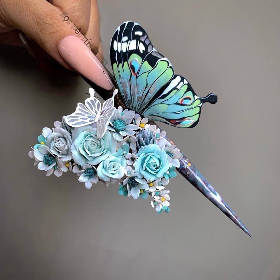 Long, embellished nail art: turquoise butterfly and flowers adorn a sharp, angled nail.