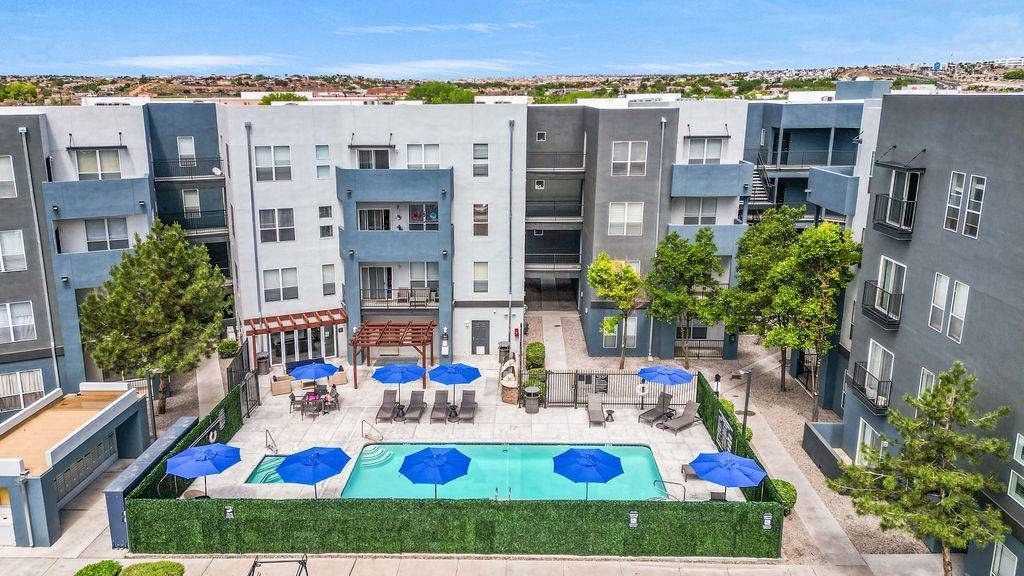 Communal pool area with blue umbrellas, lounge chairs, and modern apartment buildings around.