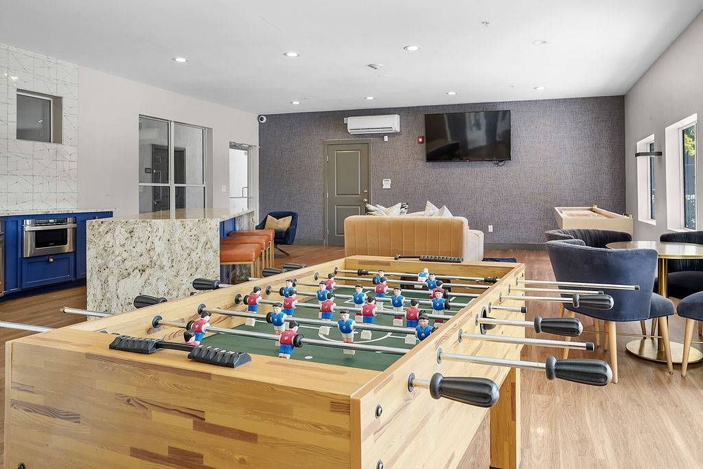 Interior of a modern apartment community lounge with a foosball table, seating, and a TV.