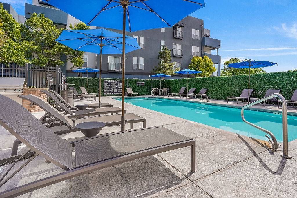 Outdoor pool area with lounge chairs, blue umbrellas, and a surrounding hedge.