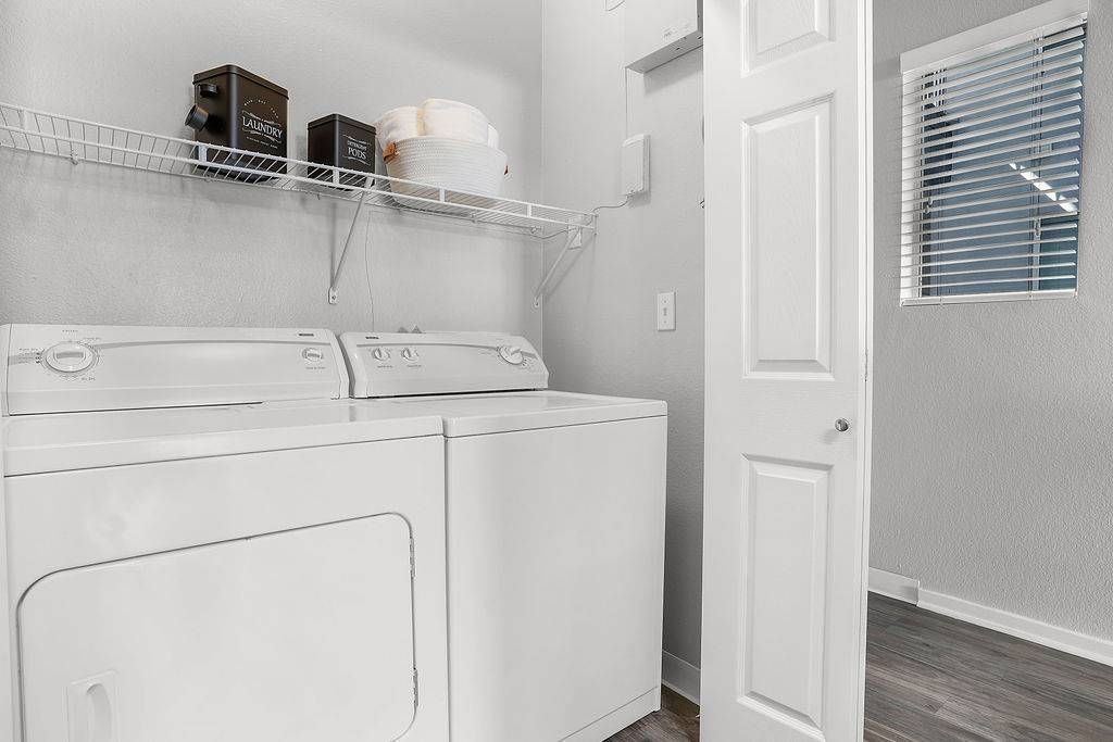 White top-loading washer and dryer side-by-side in a laundry room with a wire shelf above.