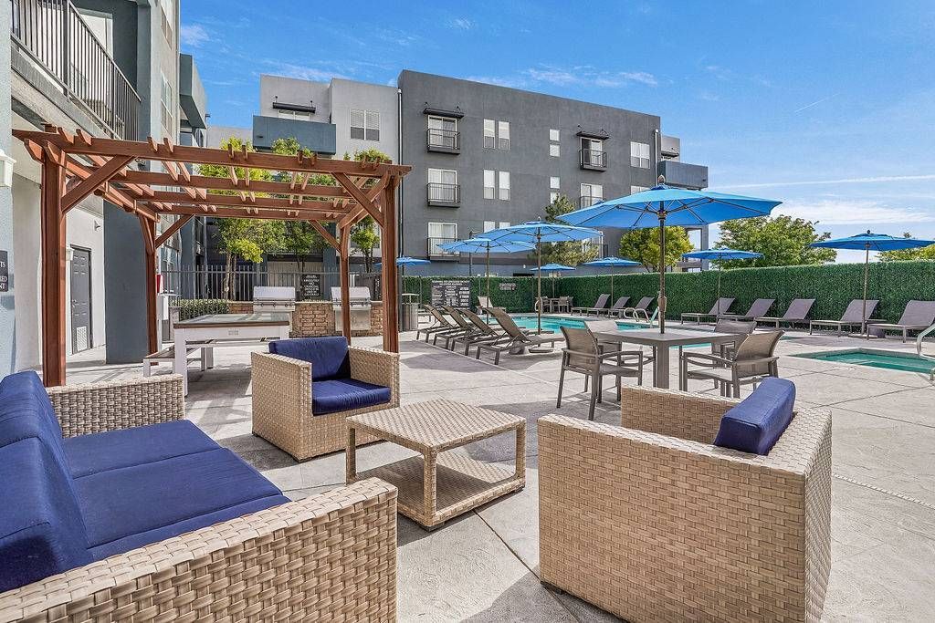 Outdoor apartment community pool deck with wicker seating, lounge chairs, umbrellas, and a pergola.
