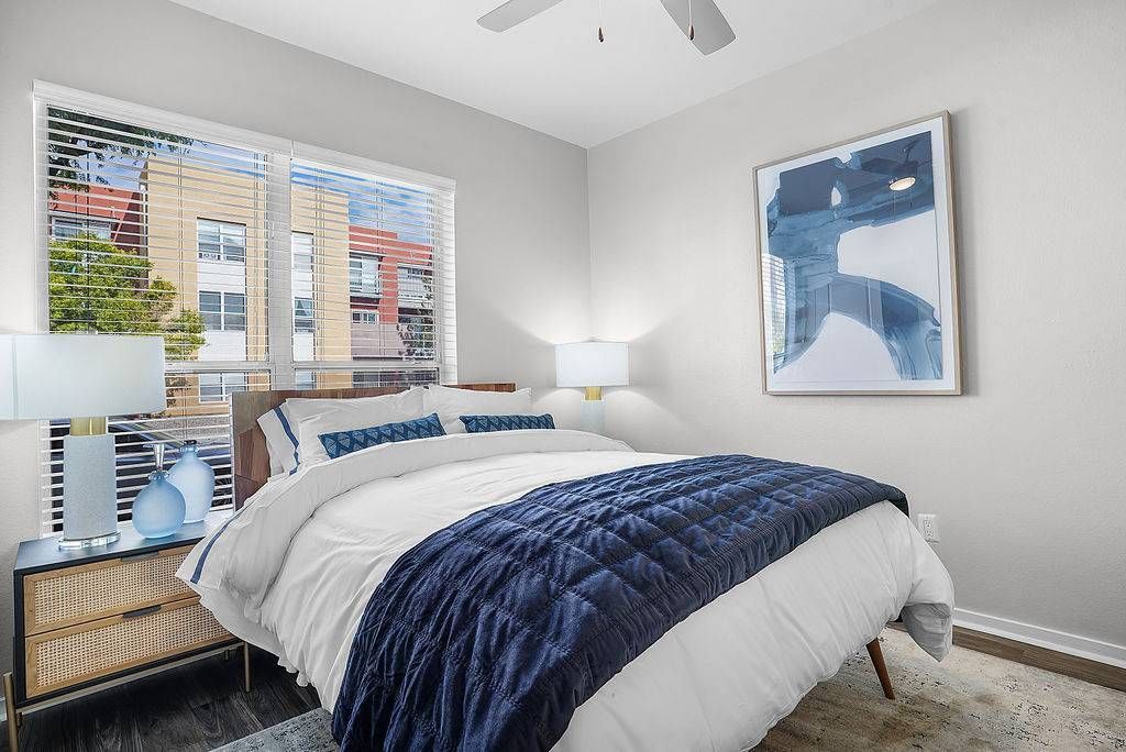 Bedroom in apartment with a queen bed, navy quilt, two nightstands, lamps, and a large window with blinds.
