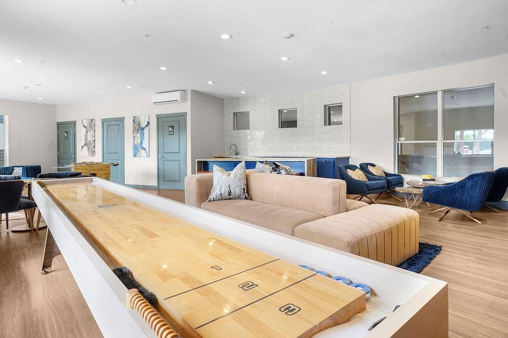 Indoor community lounge with beige sectional, blue chairs, and a long shuffleboard table.
