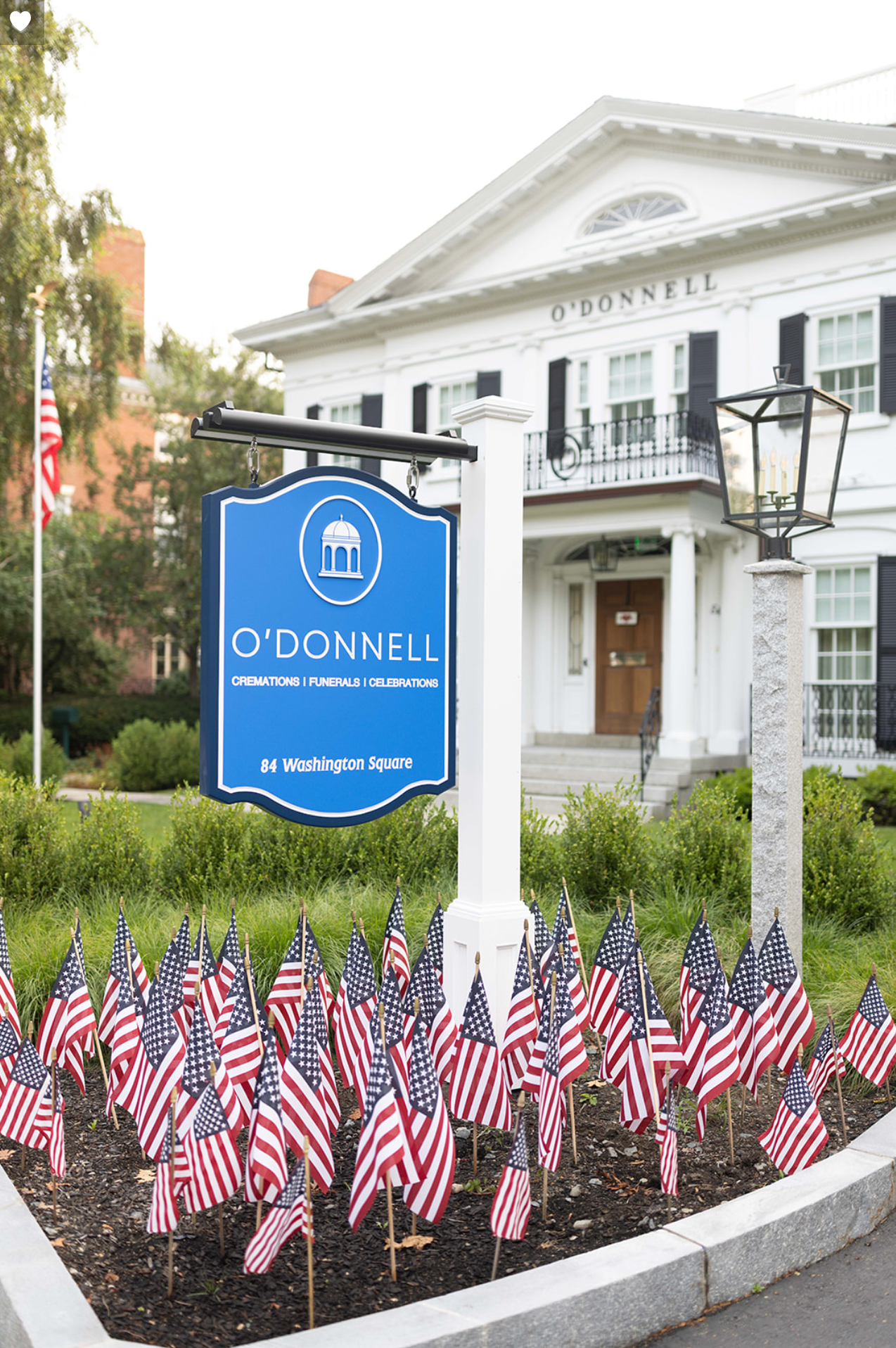 Sign for O'Donnell Funeral Home in front of white building with American flags planted in the ground.