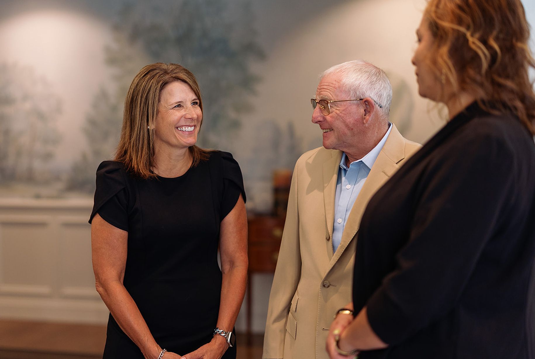 Three people conversing indoors; a woman in black smiles, facing a man in a tan jacket, and another woman in black.