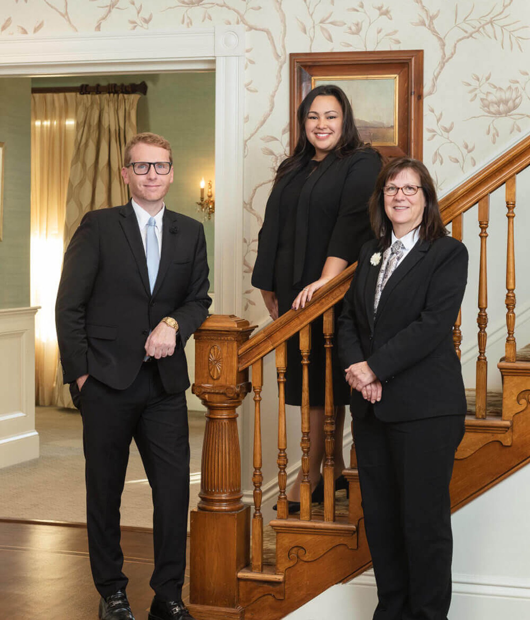 Three people in suits on a staircase. A man on the left and two women. Ornate wood staircase and wallpaper in background.