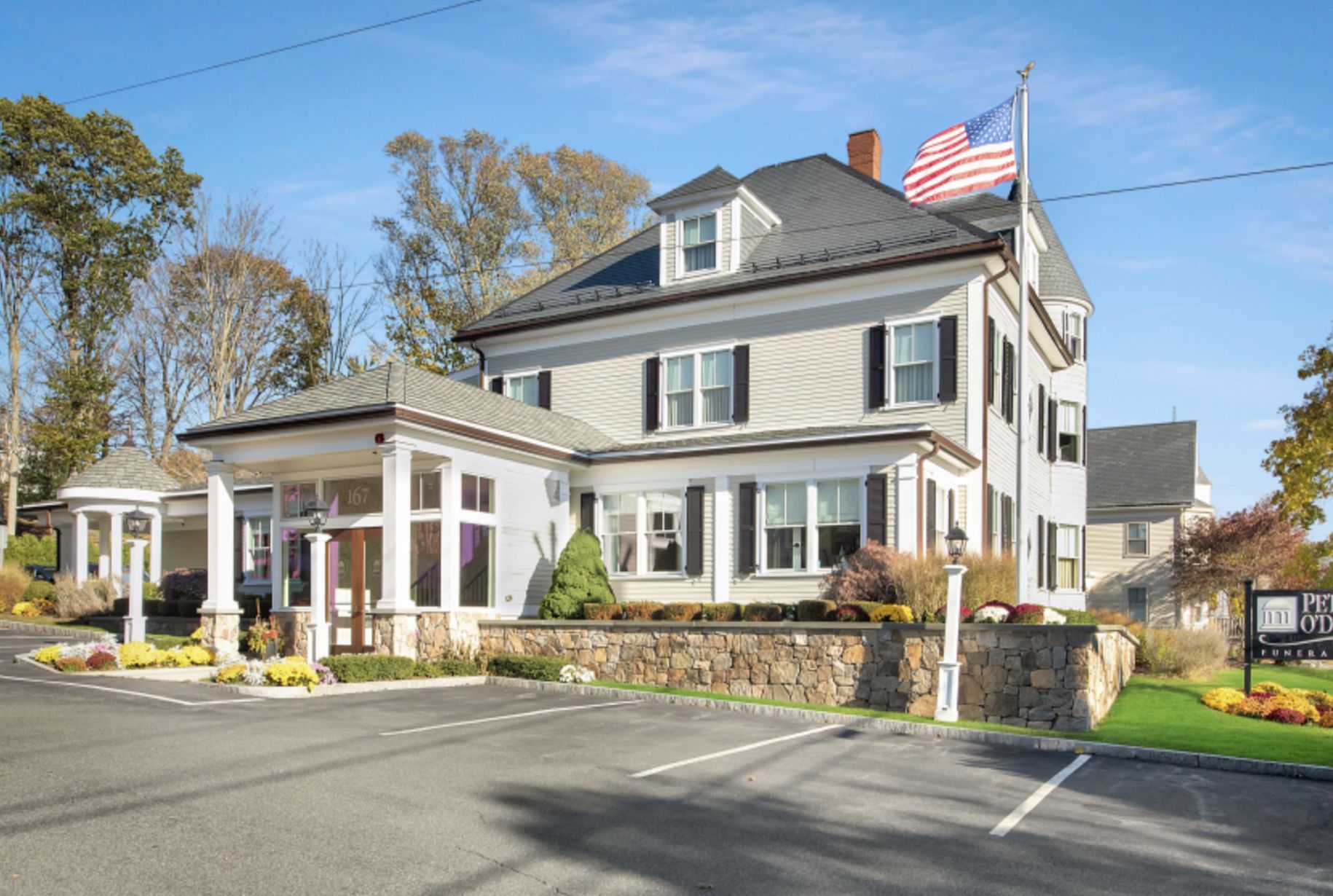 Three-story building with a light gray facade, American flag, and stone wall; a parking lot and grass lawn are in view.