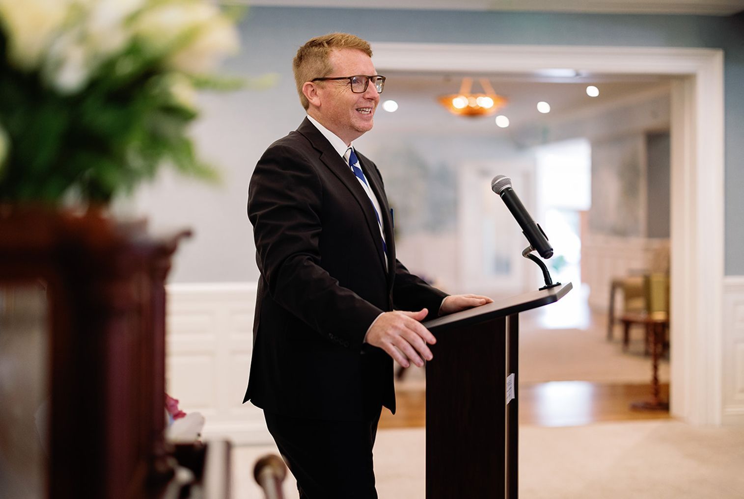 Man in a suit speaks at a podium with a microphone, smiling, inside a well-lit room.