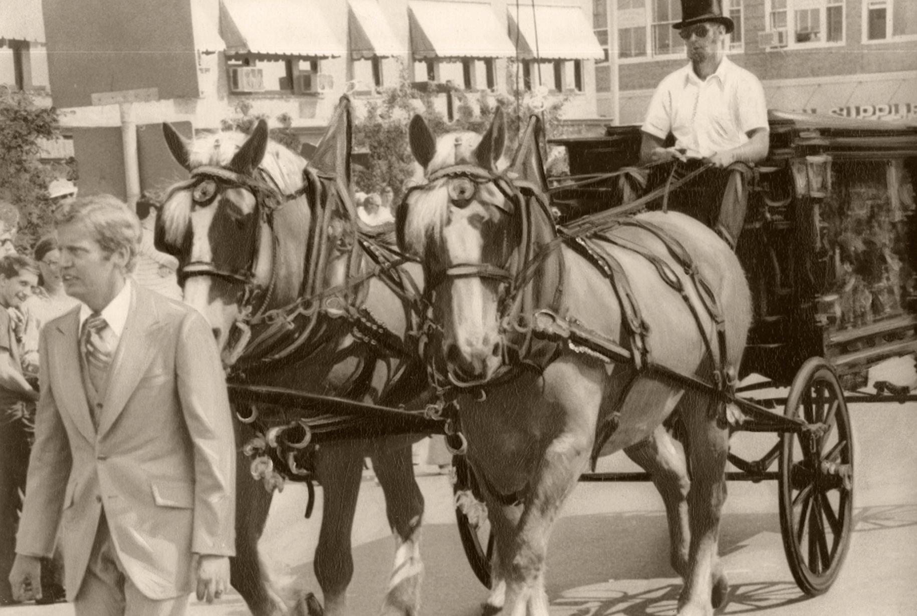 Man in suit walking next to horse-drawn carriage. Driver in top hat. Buildings in background.