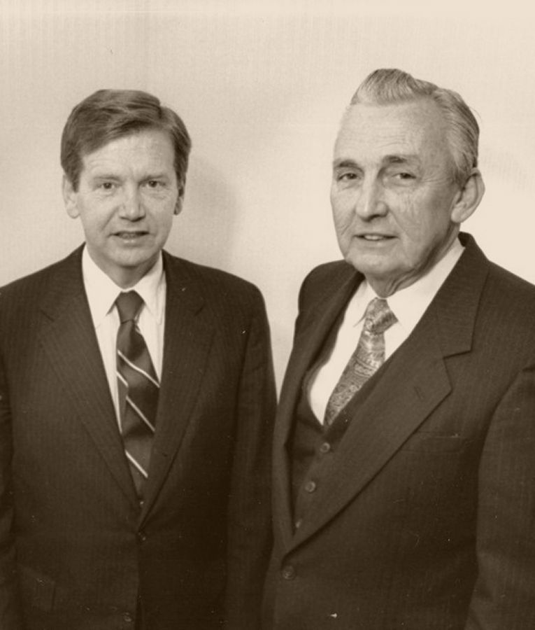 Two men in suits, smiling. One has a patterned tie, the other a plain tie and vest.