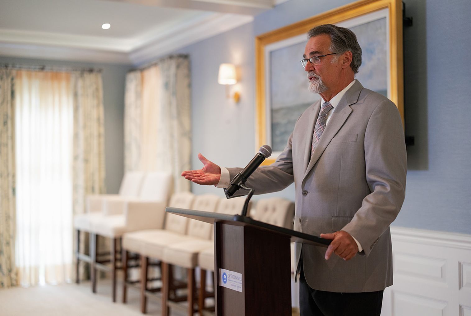 Man in light suit speaks at podium, gesturing with hand, in front of chairs and window.