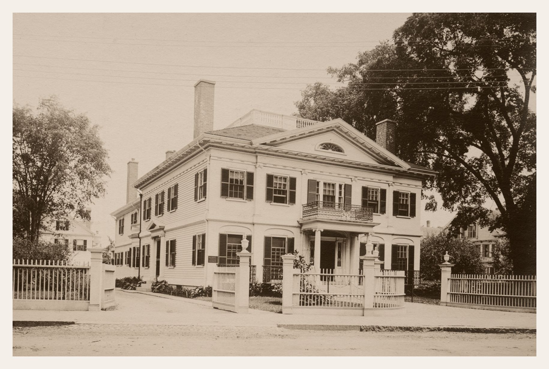 A white, two-story house with black shutters, a porch, and a wooden fence. Trees flank the house.
