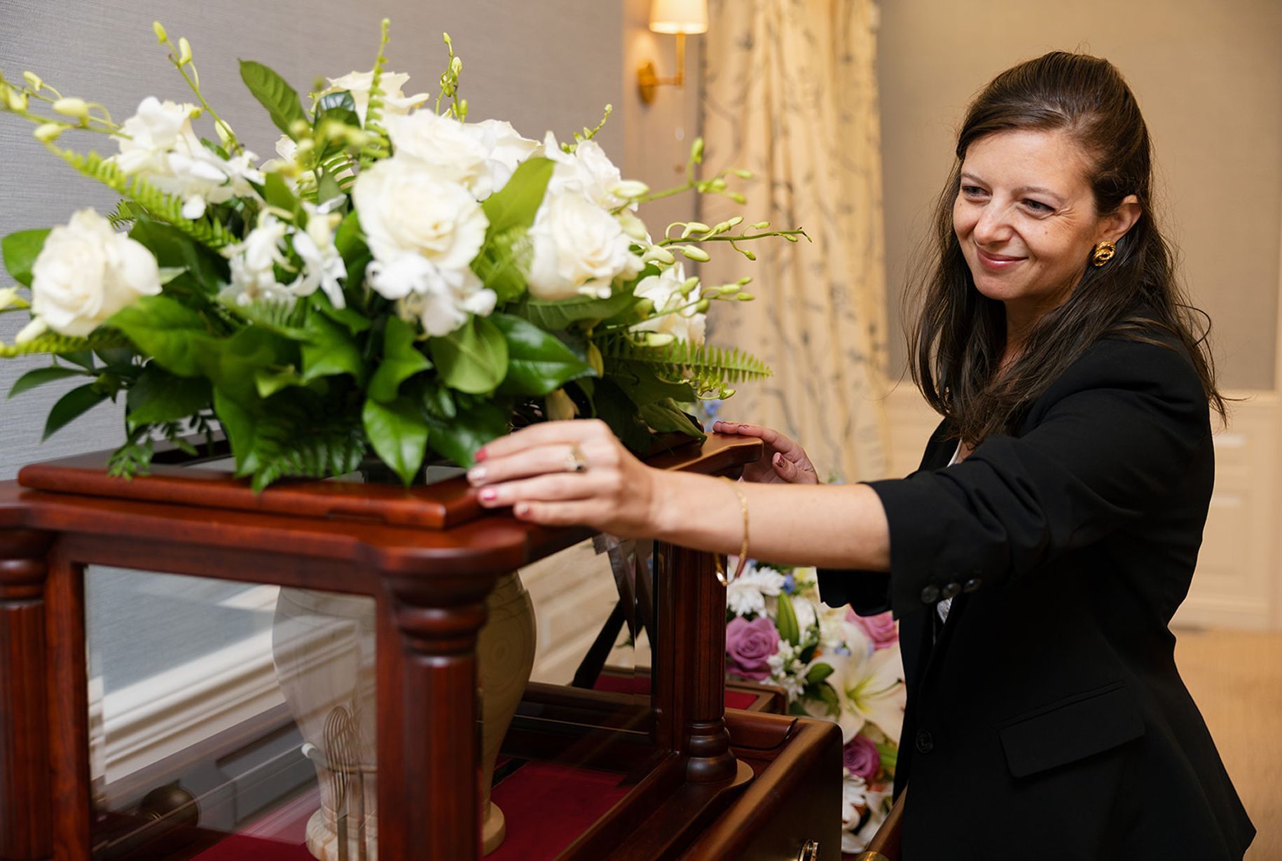 Woman arranging flowers near a display case in a room with neutral walls, soft lighting.