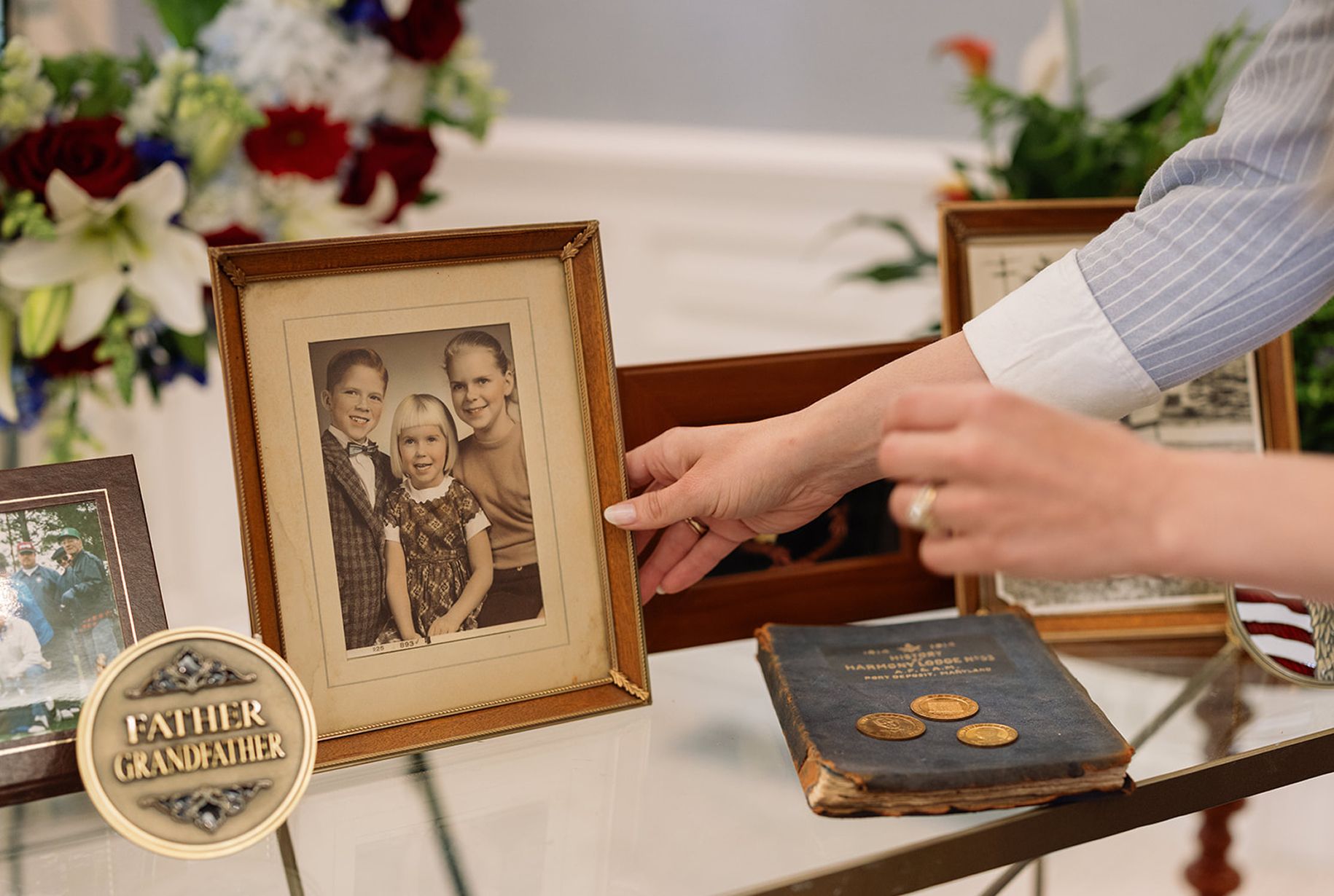 Hands arranging framed photos and a book on a reflective surface, with floral arrangements in the background.
