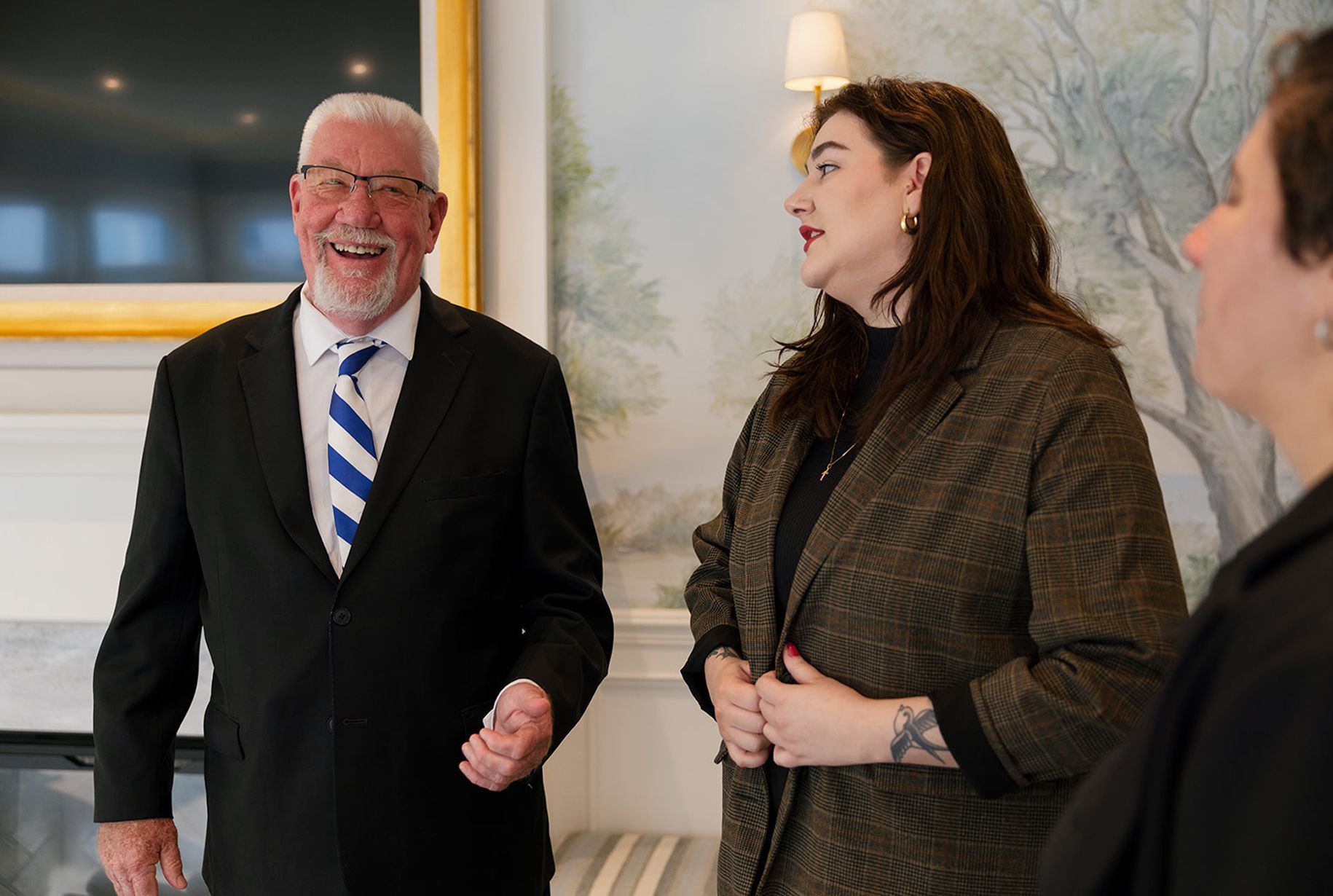 Man in suit laughs with two women in a room. One woman talks. Light-colored walls.