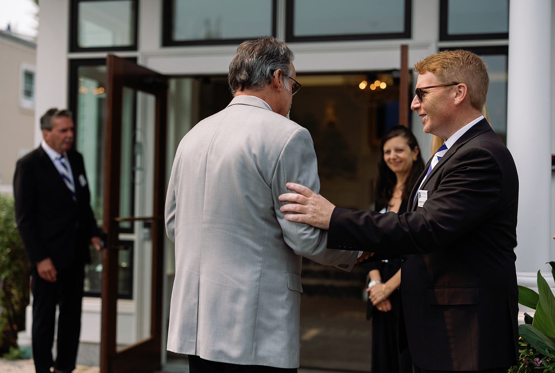 Men greet outside a building. One man in a suit, pats another on the back. A woman and another man stand in the background.
